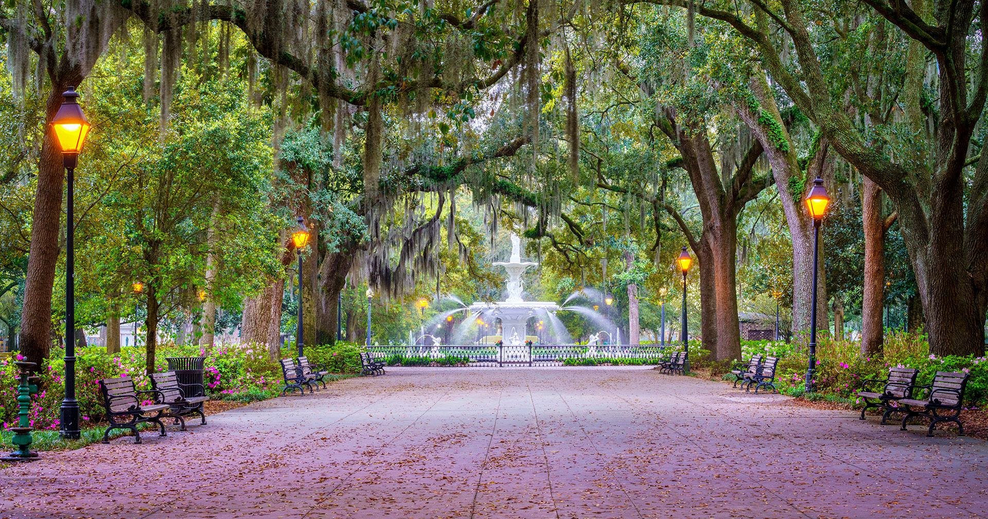 The photo depicts a serene park setting with a long pathway lined by street lamps, trees, and benches, leading towards a fountain in the distance.